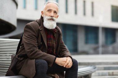 Gri saçlı ve uzun sakallı düşünceli bir adam sokakta bankta oturuyor, kalın bir ceket giyiyor, deri çanta giyiyor, iş merkezinin yanında dinleniyor, yan yana bakıyor, boşluğu kopyalıyor.