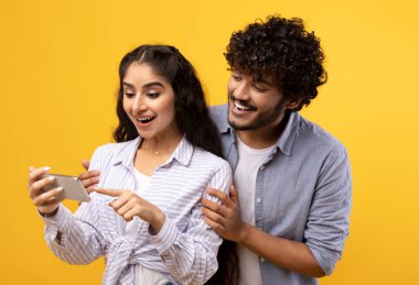 Heyecanlı Hintli çift akıllı telefon ekranına bakıyor, yeni bir uygulama kullanıyor ya da havalı haberler okuyor, sarı stüdyo arkaplanının başında duruyor. Mutlu adam ve kadın cep telefonu kullanıyor, erkek kız arkadaşını kucaklıyor.