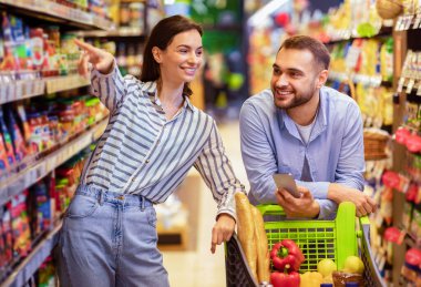 Bunu istiyorum. Milenyum çifti birlikte market alışverişi yapıyor, erkek arabaya yaslanıyor ve cep telefonu kullanıyor, kadın rafı işaret ediyor, aile yemekleri seçiyor ve marketten erzak alıyor.
