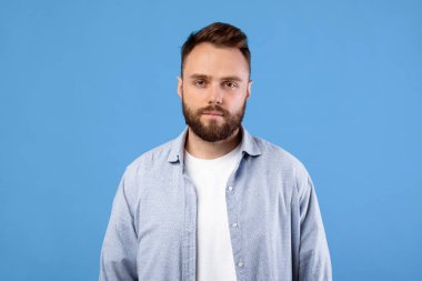 Mavi stüdyo arka planında kameraya bakan ciddi bir gencin portresi. Koyu renk saçlı ve sakallı, kendinden emin ve odaklanmış, başarılı bir milenyum erkeği.