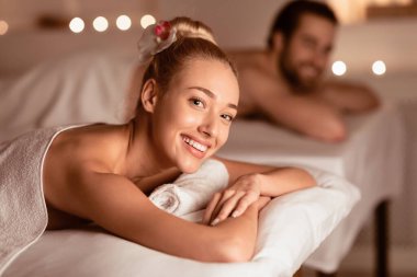 Çift Masajı. Kameraya gülümseyen mutlu kadın lüks spa salonundaki çift için rahatlatıcı spa prosedürünün tadını çıkarıyor. Romantik Güzellik Randevusu Konsepti. Seçici Odaklanma