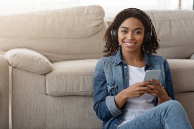 Evde kal. Kablosuz kulaklık takan, akıllı telefondan müzik dinleyen, karantinada dinlenen gülümseyen Afrikalı kadın.