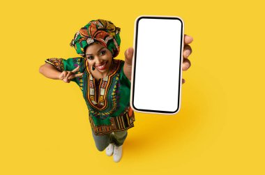 Beyaz beyaz ekranlı, güzel siyah kadın eli ile akıllı telefon, ulusal Afrika kostümü giyen mutlu genç bayan harika bir online anlaşma ve barış jesti gösteriyor, sarı stüdyo arka planı, model, üst görünüm