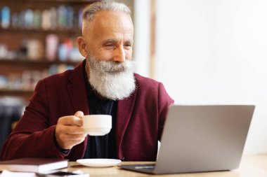 Gri saçlı, kahve içen ve kafeteryada dizüstü bilgisayar kullanan yaşlı bir adam. Şık, şatafatlı, bordo ceketli iş adamı internette çalışırken çayının keyfini çıkarıyor.
