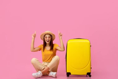 Yazlık şapkalı ve güneş gözlüklü duygusal genç kadın bavullarıyla yerde oturuyor ve fotokopi alanını işaret ediyor, pembe stüdyo arka planı. Yaz kıyafetleri içindeki mutlu bayan heyecan verici seyahat anlaşmaları gösteriyor.