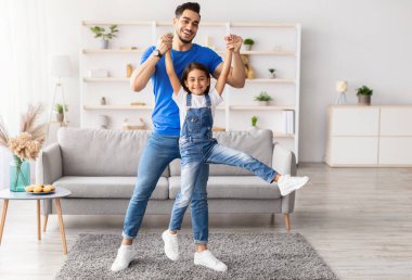 Sevgi dolu bir aile. Tam vücut uzunluğunda gülümseyen bir baba, küçük tatlı kızıyla birlikte oturma odasında boş zaman geçirir. Ellerini tutar, kaldırır, eğlenir ve zıplar.