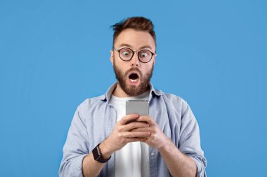 Şok olmuş Kafkasyalı bir adam mavi stüdyo arka planı ve panorama üzerinden cep telefonuna bakıyor. Mobil cihazlı genç adam inanılmaz haberler öğreniyor, reklam satışına hayran kalıyor.