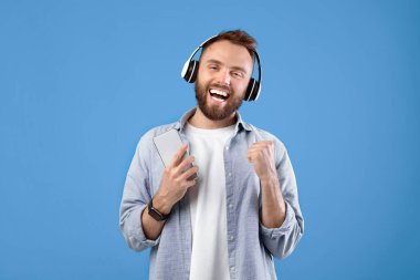 Cep telefonuyla kulaklıkla müzik dinleyen, mavi stüdyo arka planında en sevdiği şarkıyı söyleyen yakışıklı beyaz adam. Popüler melodileri seven pozitif genç adam