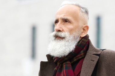 Sıcak kıyafetler içinde şık yaşlı bir adam ofise gidiyor, kahve içiyor, deri çanta taşıyor, yan bakıyor. Yaşlı gri saçlı adam caddede yürüyor, kahvesini içiyor, iş merkezi arkada.