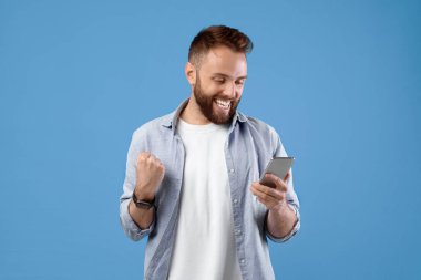 Akıllı telefon kullanan mutlu sakallı bir gencin portresi mavi stüdyo arka planında birinci sınıf bir jest yapıyor. Heyecanlı adam zaferi kutluyor, başarıya ulaşıyor, EVET işareti yapıyor