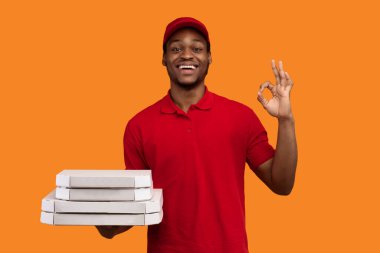 Fast Service ve salgın sırasında en iyi yemek teslimatı. Kırmızı üniformalı ve şapkalı bir siyah kurye restorandan pizza kutuları taşıyor ve turuncu arka planda izole edilmiş işaret işareti gösteriyor.