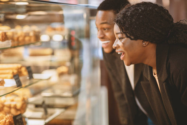 Couple together choosing sweets. Surprised happy millennial woman and man standing in front of glass showcase with pastries and cakes in modern grocery store, side view, close up, free space