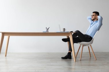 Ara veriyoruz. Sırıtkan Arap bir gencin masada oturup dizüstü bilgisayarı kullanmasının profil yan görüntüsü. Mutlu milenyum erkeğinin iş yerinde arkasını yaslayıp bilgisayar ekranına bakması.