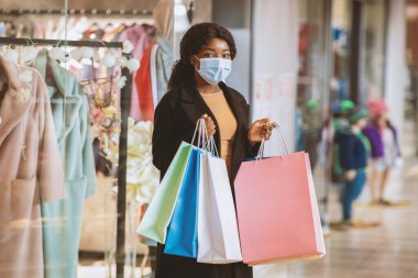 Güzel Afro-Amerikalı bir kadının portresi hafta sonunu alışveriş merkezinde geçiriyor. Covid-19 'daki sosyal uzaklık sırasında. Koruyucu maskeli kadın butik vitrininin yanında renkli çantalar tutuyor.