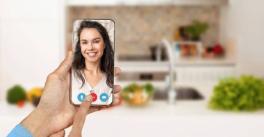 Bir insan, mutfak ortamında video görüşmesi yapmak için akıllı telefon kullanıyor. Ekranda gülümseyen bir katılımcıyı gösteren dostane bir jest var..