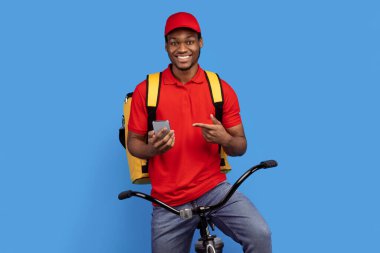 Kurye Hizmeti Başvurusu. Akıllı telefon kullanan, kırmızı üniformalı, şapkalı, termal çantalı, bisikletli, mavi stüdyo arka planında izole edilmiş gülümseyen siyah bir teslimatçı.