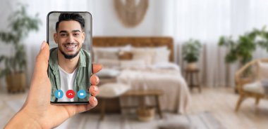 Bir kişi, rahat evlerinden gelen bir video görüşmesi sırasında gülümseyen bir şahsı gösteren bir akıllı telefon tutuyor. Arka planda sıcak, davetkar bir yatak odası ortamı var..