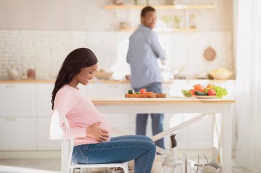 Hamilelik sırasında sağlıklı beslenme. Güzel hamile kadın karnına dokunurken kocası mutfakta yemek pişiriyor, fotokopi odası. Siyah aileyi sevmek, bebek beklerken dengeli yemek yapmak.