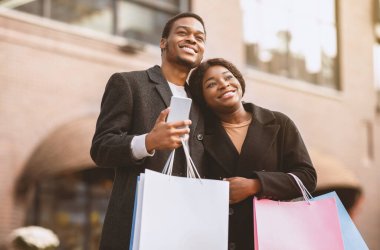 Kara Cuma 'da harika alışveriş ve satış reklamı. Renkli çantalı mutlu Afro-Amerikan erkek ve kadın sarılıyor ve inanılmaz bir şeye bakıyor. Bir erkek akıllı telefonu dükkanın yakınında tutuyor.