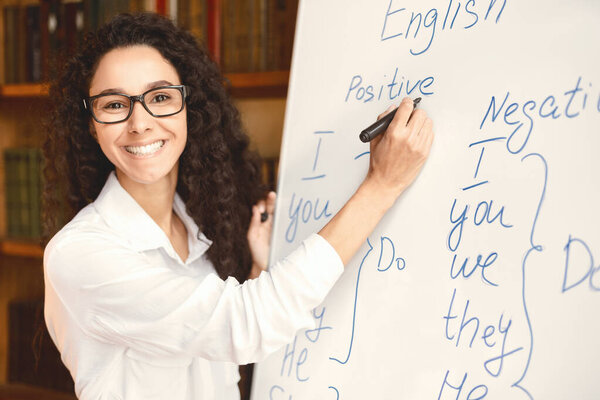 Lecture Concept. Portrait of positive female English teacher standing at board, writing grammar rules with marker. Smiling tutor in eyeglasses looking at camera, explaining new theme of the lesson
