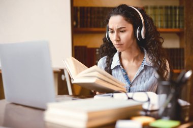 Uzaklık Eğitimi. Kablosuz kulaklık takmış, kağıt kitap okuyan, dizüstü bilgisayarla masada oturan ciddi bir kadının portresi. Ev ödevi yapan bir bayan, evde uzaktan ders çalışan. E-öğrenme