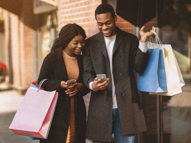 Mobil cihazda sezonluk satışla ilgili bir mesaj var. Gülümseyen Afro-Amerikan karı ve koca paltolu renkli alışveriş paketlerini tutuyor ve butiğin yanındaki akıllı telefona bakıyor, fotokopi çekiyor.