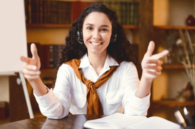 Video konferansı. Gülümseyen genç bir kadının internet araması ya da sanal görüşme yaparken ön portresi, evdeki ofiste ya da kütüphanede otururken kamerayla konuşmak, kablosuz kulaklık takmak, açıklama yapmak.