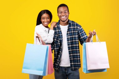 Alışveriş için boş zaman, kara cuma ve reklam alanı. Neşeli milenyum Afrikalı Amerikalı erkek ve kadın öğrenci birçok renkli alışveriş torbası sergiliyor, sarı arka planda izole edilmiş, stüdyo çekimi