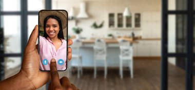 Bir kişi video görüşmesi sırasında neşeli bir kişiyi gösteren bir akıllı telefon tutuyor. Arka planda rahat bir ev atmosferini gösteren rahat bir mutfak var..