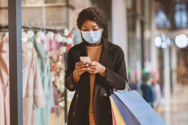 Şehir merkezindeki covid-19 salgınında sosyal mesafe ve yeni normallik. Koruyucu maskeli ve ceketli genç Afrikalı Amerikalı kadın renkli paketler taşıyor ve vitrindeki akıllı telefona bakıyor.