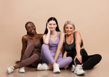 Üç farklı etnik kadının portresi spor kıyafetleri içinde mutlu vücut pozları, farklı boyutlarda ve bej zeminde oturan ve kameraya gülümseyen kadınlar.