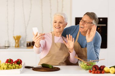 Uzaktan iletişim için cep telefonu kullanan neşeli yaşlı çift modern mutfaktaki cep telefonuna el sallıyor. Uzaktan Aile Arayan Yaşlılar Klibi