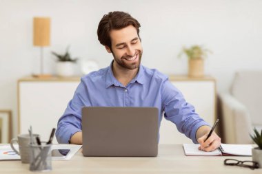 Dizüstü bilgisayarda çalışan neşeli iş adamı modern ofiste işyerinde oturup notlar alıyor. Seçici Odaklanma. Başarılı İş Kariyeri ve Girişimcilik Konsepti.