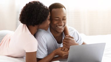 Romantik siyah erkek ve kadın karı koca, evde yatakta dinlenirken modern dizüstü bilgisayar kullanırken kucaklaşıyorlar, fotoğraf izliyorlar ya da birlikte web sörfü yapıyorlar, yatak odasında tembelce hafta sonları geçiriyorlar, uzay kopyaları yapıyorlar.