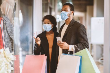 Satış ve Kara Cuma alışverişi. Mutlu, Afro-Amerikalı, koruyucu maskeli bir çift alışveriş merkezinde yürür, renkli çantaları ellerinde tutar, akıllı telefonu kontrol için kullanır ve vitrinlere çıkarlar.