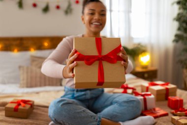 Noel hediyesi konsepti. Heyecanlı Afrikalı Amerikalı kadın hediye kutusunu kameraya uzatıyor ve gülümsüyor, seçici odaklanıyor. Çeşitli hediye kutularıyla dolu yatakta oturan neşeli siyahi kadın.
