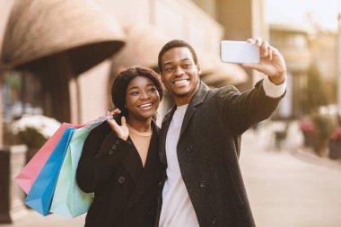 Kasım ayında Kara Cuma 'da büyük mevsimsel indirimde alışveriş yaparken selfie. Gülümseyen Afrikalı genç Amerikalı erkek ve kadın akıllı telefona bakıyor ve şehir merkezinin yakınındaki sokakta fotoğraf çekiyor.