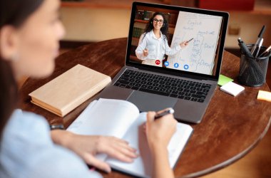 E-öğrenme kavramı. Masada oturan, deftere yazan, not alan, dizüstü bilgisayar kullanan, İngilizce öğretmeniyle çevrimiçi ders alan kız öğrencinin omzunun üstünden. Uzak çalışma