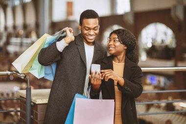Kara Cuma ve çevrimiçi alışveriş ve nakit iade için mobil uygulama. Renkli paketleri olan neşeli Afro-Amerikalı genç kadın, şehir merkezindeki indirimle ilgili mesajlı akıllı telefonu erkeklere gösteriyor.
