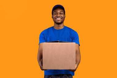 Teslimat servisi konsepti. Mavi şapkalı ve tişörtlü gülümseyen siyah postacı karton kutu tutuyor, kameraya gösteriyor ve ona veriyor, kopyalama alanı. Turuncu stüdyo arka planında duran postacı.