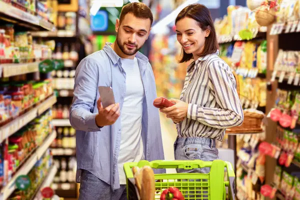 Süpermarkette alışveriş yapan genç bir ailenin portresi. Gülümseyen Çift Kapalı Kapılar 'da Gıda Ürünleri Taraması Yapıyor. Müşteriler Malzeme Alırlar, Fiyatı Karşılaştırırlar