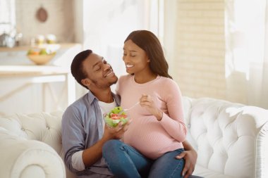 Hamilelik konseptinde dengeli diyet. Yakışıklı siyahi adam, sevgili müstakbel karısına içeride lezzetli sebze salatası veriyor. Pozitif siyah çift bebek beklerken sağlıklı besleniyorlar.