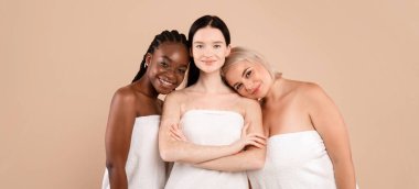 Farklı ten ve vücut tiplerine sahip mutlu çok ırklı kadınlar bej rengi stüdyo arka planında duran banyo havluları giyerler, spa tedavisinden sonra poz veren çekici genç ırklar arası kadınlar, kopyalama alanı.