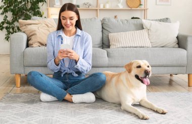 Yaşam tarzı kavramı. Saf köpeğiyle halıya oturmuş akıllı telefon kullanan gülümseyen kadının portresi. Bayan evde vakit geçiriyor, sosyal medyada sohbet ediyor, SMS atıyor ya da mesaj okuyor.