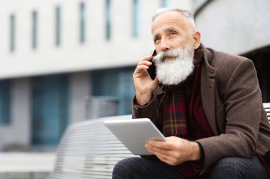 Uzun sakallı, yaşlı, elinde dijital tabletle bankta oturan, cep telefonuyla konuşan, sokakta dinlenirken iş görüşmesi yapan, boş alana bakan gri saçlı bir adam.