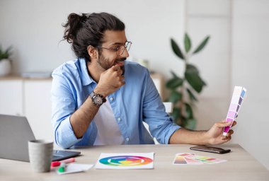 Genç Hintli erkek tasarımcı renk örneklerini tutuyor ve inceliyor, yaratıcı ofiste masa başında çalışıyor, iç tasarımı seçiyor, yeni proje için renk gama 'sını kontrol eden şık batılı adam, serbest alan