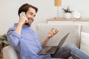Evdeki koltukta dizüstü bilgisayarla oturup konuşan neşeli adam. Modern İletişim ve Yaşam Tarzı. Mutlu adam cep telefonuyla konuşuyor ve içeride gülüyor.
