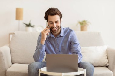 İş konuşması. Cep telefonuyla konuşan, bilgisayarını kullanan evden uzakta çalışan, koltukta oturan bir adam. Freelancer bilgisayarda çalışırken telefonla konuşuyor. Serbest ve Mesafeli Çalışma Konsepti