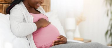 Neşeli siyah hamile kadın evde yatakta dinleniyor, göbeğine dokunuyor ve gülümsüyor, büyük göbeğine bakıyor, hamileliğinin tadını çıkarıyor ve mutlu beklenti zamanı, yatak odasında dinleniyor, yan manzara, boş alan...