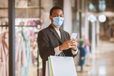 Coronavirus ve sosyal mesafe süresince mobil uygulamalarda online sipariş ve alımlar. Milenyum Afrika kökenli koruyucu maskeli bir Amerikalı akıllı telefonuyla yazı yazıyor ve elinde kağıt alışveriş torbaları tutuyor.